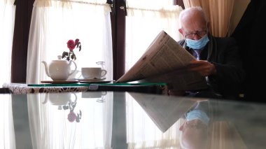 Gri saçlı, gözlüklü ve siyah ceketli yaşlı bir adam pencerenin kenarında bir sandalyede oturuyor, yeni bir gazete okuyor, başka tarafa bakıyor, gazete okuyor. Gerçek zamanlı. 4k video