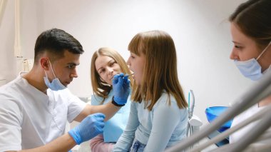 Bir dişçi ve asistanları kızıl saçlı genç bir kızın ağız boşluğunu inceliyor. Gençlerde diş tedavisi uygulanıyor. Profesyonel diş tedavisi