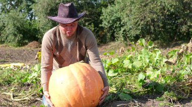 Farmer young man in work clothes and hat on his head holding a pumpkin in his hands, harvest season. organically pure vegetables from the garden