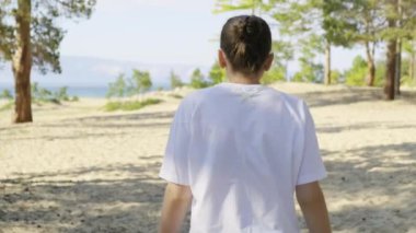 Beyaz tişörtlü bir kız güneşli bir yaz gününde Baykal Gölü 'ndeki Olkhon Adası sahilinde yürüyor. Yavaş çekim, geri çekim, takip çekimi.