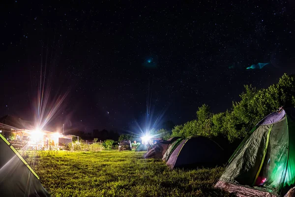 Night camping under starry sky. Glowing tourist tent, under beautiful evening sky full of stars and Milky way, city lights on background. Outdoor lifestyle concept.
