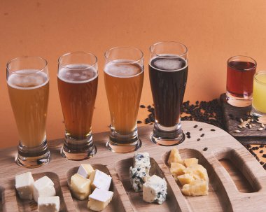Turuncu arka planda, ahşap bir masada farklı türde soğuk bira. Bira, buğday, koyu hafif bira, bira konsepti. Alkolik içki afişi. Peynirin farklı çeşitleri olan peynir tabağı.