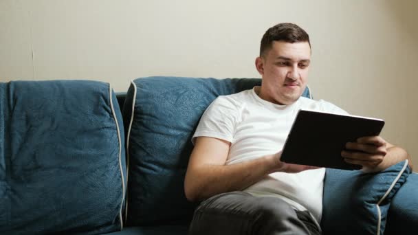 Een 30-jarige man met een Europees uiterlijk zit op een donkerblauwe bank en werkt op een tablet. Guy glimlacht naar het tabletscherm. Stockvideo's