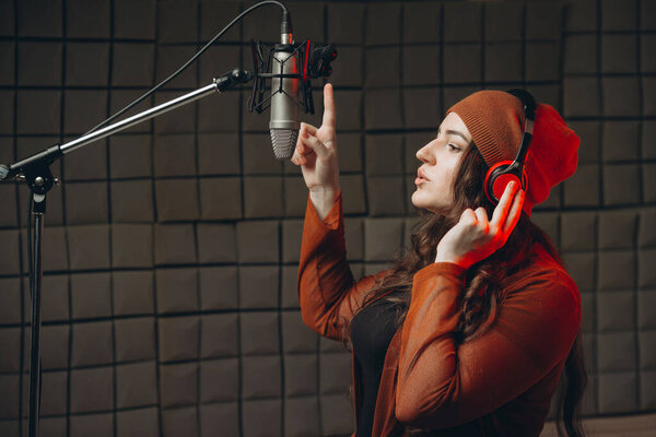 Young woman in headphones emotionally singing in microphone in the sound recording studio. Star, talent, preparing show, studio recording. Soundproof room for professional recording vocal