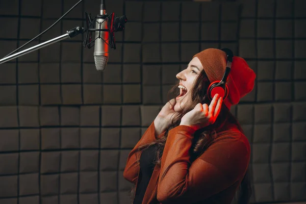 Young woman in headphones emotionally singing in microphone in the sound recording studio. Star, talent, preparing show, studio recording. Soundproof room for professional recording vocal