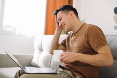 Adam kanepede oturup kahve içiyor ve lapto kullanıyor.