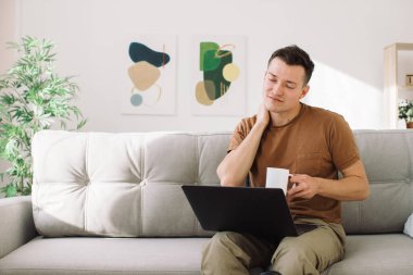 Oturma odasındaki kanepede oturan stresli genç beyaz adam, dizüstü bilgisayar kullanıyor, serbest çalışan, uzaktan çalışan, baş ağrısı çeken, dinlenmesi gereken biri.