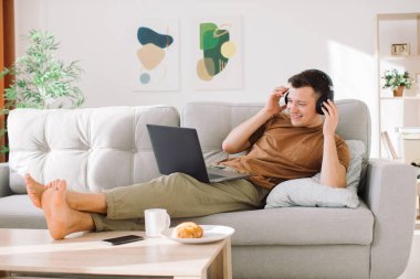 Kablosuz kulaklıklı rahat genç adam minimalist oturma odasında kanepede uzanıyor ve çevrimiçi çalışırken dizüstü bilgisayar kullanıyor.