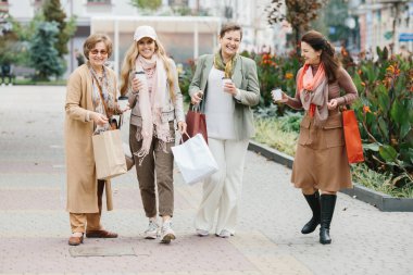 Bir grup gülümseyen yetişkin kadın şehirde yürürken ellerinde poşetlerle dolaşıyorlar.