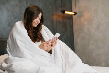 Pretty girl with dark hair having relaxation, holding mobile phone, sitting on bed wrapped with blanket after awakening.