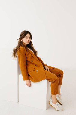 Beyaz arka planda küp üzerinde oturan modaya uygun pastel kıyafetli genç kadın.