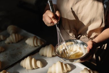 Female baker smears raw croissants with a brush in the yolk, the cooking process.