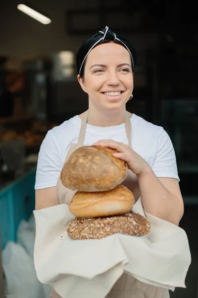 Shot of an cheerful female baker smiling to the camera standing near the showcase. Female baker or saleswoman in her bakery selling fresh bread, pastries and bakery products.