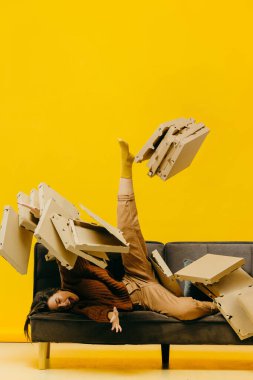 Yaratıcı pizza teslimatı kavramı. Kanepedeki kız pizza kutularıyla kaplı. Pizza kutularından oluşan bir çığ..