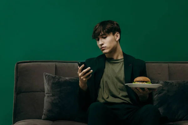 A young man in a green stylish suit is eating a burger while sitting on a sofa on a green background. Fast food concept.