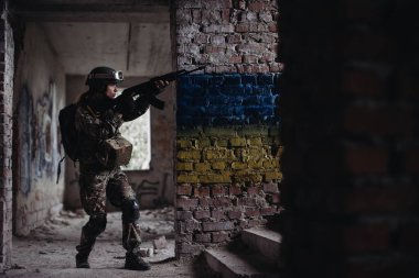 Ukrayna bayrağının arkasında duran askeri kadın asker. Ukrayna bayrağı bir tuğla duvara boyanmış. Elinde silah olan bir Ukraynalı savunucu..