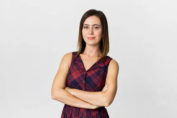 Çekici bakışlı, kollarını kavuşturmuş, renkli elbiseler giymiş, beyaz boş duvara karşı poz veren pozitif bir kadın.