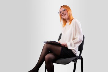 Beyaz bluzlu bir kız, elinde dosyayla, neşeli bir şekilde gülüyor, gri bir arka planda.