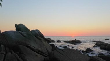 Gün batımında adanın yanındaki ağaçların üzerinde güneşin ufka doğru batışını gösteren hava manzarası. Adanın etrafındaki taşlarla dolu kaya plajı. Okyanus zemininin üstünde günbatımı manzarası..