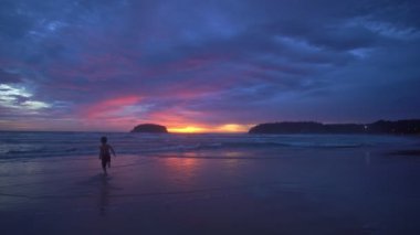 Gün batımında sahilde koşan mutlu bir çocuk. 4K UHD Mavi gökyüzü ve renkli bulutları olan güzel tropikal plaj. Dalgaların boş plajı vurduğu tropik sahil. Phuket Tayland 'da..