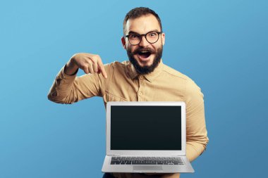 Sakallı adam bej gömlek ve gözlük takıyor. Yeni dizüstü bilgisayarı işaret ediyor.