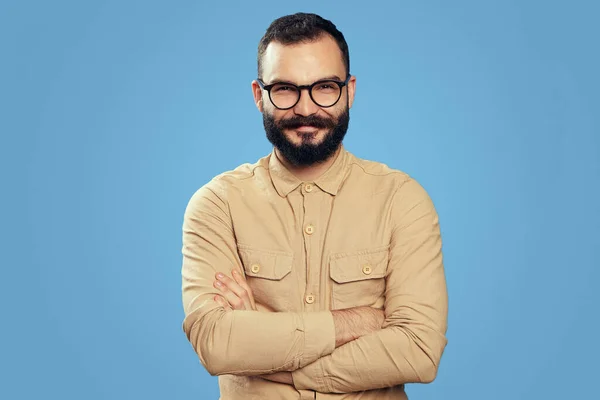 Jovem barbudo sorrindo com as mãos cruzadas, isolado sobre fundo azul — Fotografia de Stock