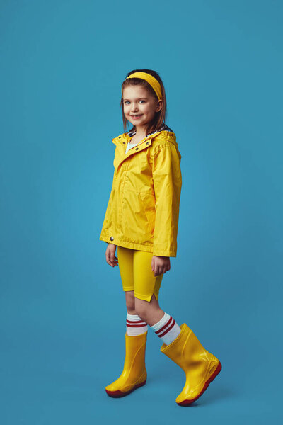 Little girl in yellow raincoat and sneakers smiles while standing against blue