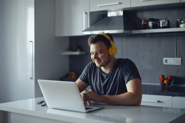 Gülümseyen serbest çalışan genç adam bilgisayarını kullanarak evde çalışıyor.