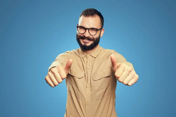 Homem com barba vestindo óculos e mostrando os polegares felizes no azul — Fotografia de Stock