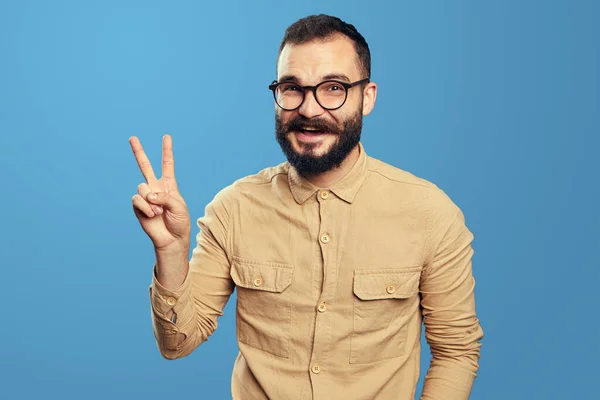 Hombre barbudo optimista sonriendo y muestra gesto de victoria sobre fondo azul — Foto de Stock