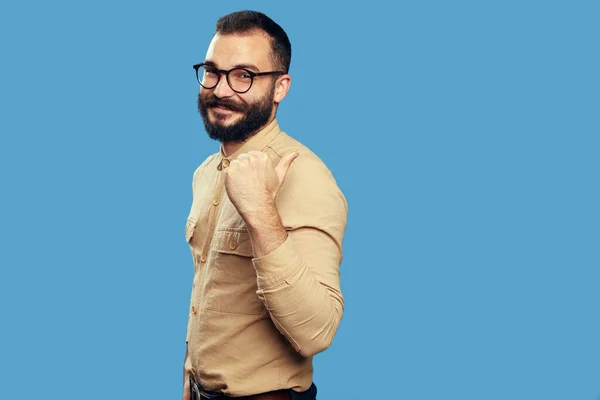 Glücklicher bärtiger Hipster zeigt zur Seite auf leeren Raum, isoliert über blauer Wand — Stockfoto