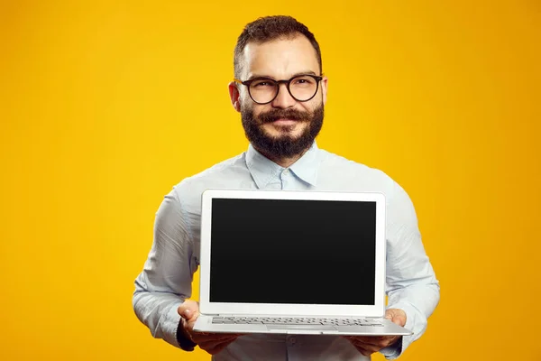 Gözlüklü ve bej gömlekli sakallı adam yeni modern laptop cihazını gösteriyor.