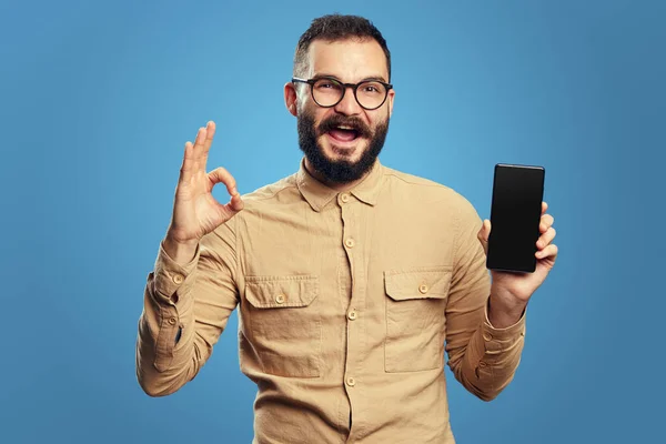 Homem barbudo jovem mostrando telefone celular tela em branco enquanto gesticulando sinal ok — Fotografia de Stock