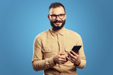 Elinde cep telefonu tutarken kameraya bakan adam, mavi arka planda izole edilmiş.