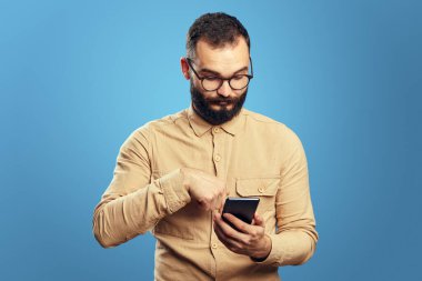 Ciddi yakışıklı beyaz adam mavi arka planda akıllı telefon kullanıyor.