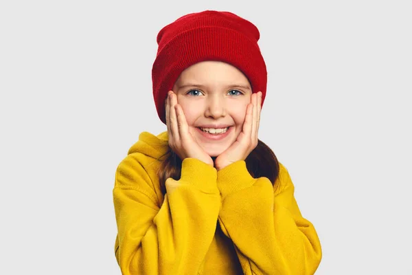 노란 후드와 빨간 모자를 쓴 사랑 스러운 작은 소녀는 뺨에 두 손을 잡고 있다 — 스톡 사진
