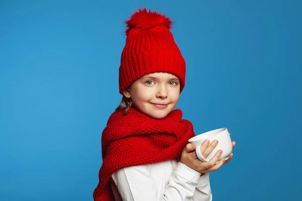 Aranyos lány kötött piros sállal és kalapban melegítő kezekkel, miközben egy csésze teát tart a kezében. — Stock Fotó