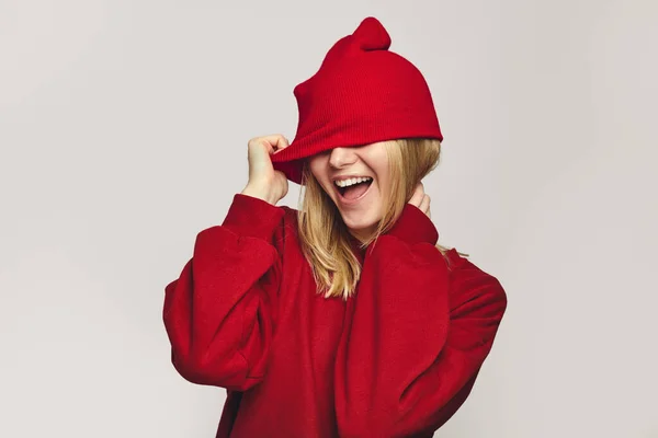 Κορίτσι με κόκκινα ρούχα που καλύπτει τα μάτια με καπέλο, χαμογελώντας στην κάμερα σε λευκό — Φωτογραφία Αρχείου