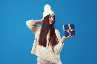 Beyaz kazaklı ve şapkalı şirin kız Noel için mavi hediye paketi taşıyor.