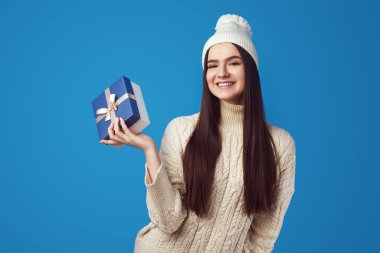 Beyaz kazak ve şapka giyen bir kız Noel için hediye kutusunu tutuyor.