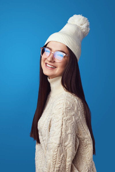 Cute girl smiles at camera, wears white hat and knitted white oversized sweater