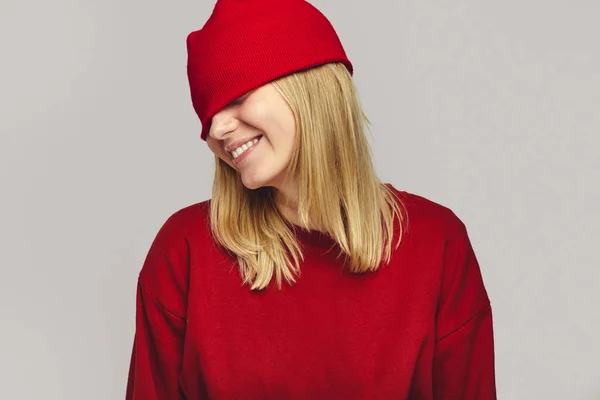 Chica en traje rojo cubre los ojos con sombrero, sonriendo ampliamente sobre fondo blanco — Foto de Stock