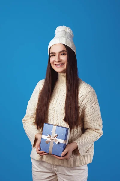 Весела мила дівчина в білому підвішеному светрі і капелюсі, тримає подарункову коробку — стокове фото