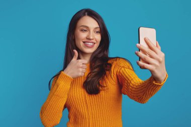 Turuncu kazaklı tatlı kız akıllı telefonuyla selfie çekerken iyi niyet gösterisi yapıyor.