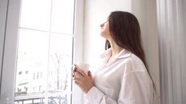 4k young woman standing near window enjoying coffee, looking at city