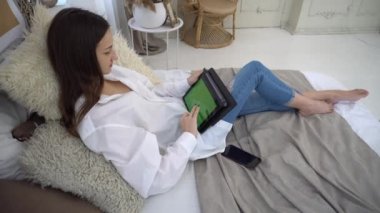 4k young woman sitting on bed and using digital tablet with green screen