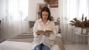 Young female sitting on bed in bedroom and reading a old memory book.
