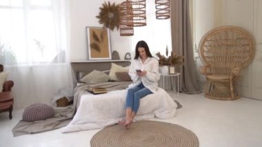 Woman in white t-shirt sitting in bed using mobile phone, typing sms message