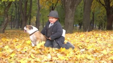 Küçük neşeli çocuk sonbahar parkında sarı yaprakların arasında köpeğin yanında oturuyor..