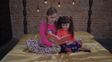 4K. Yatak odasında kitap okuyan şirin küçük kız kardeşler.